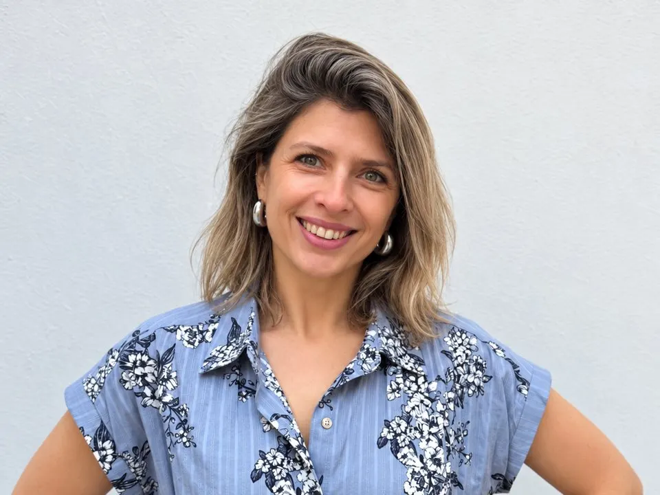 Retrato de Verónica Litvinoff, sonriente, con camisa celeste estampada frente a una pared blanca.